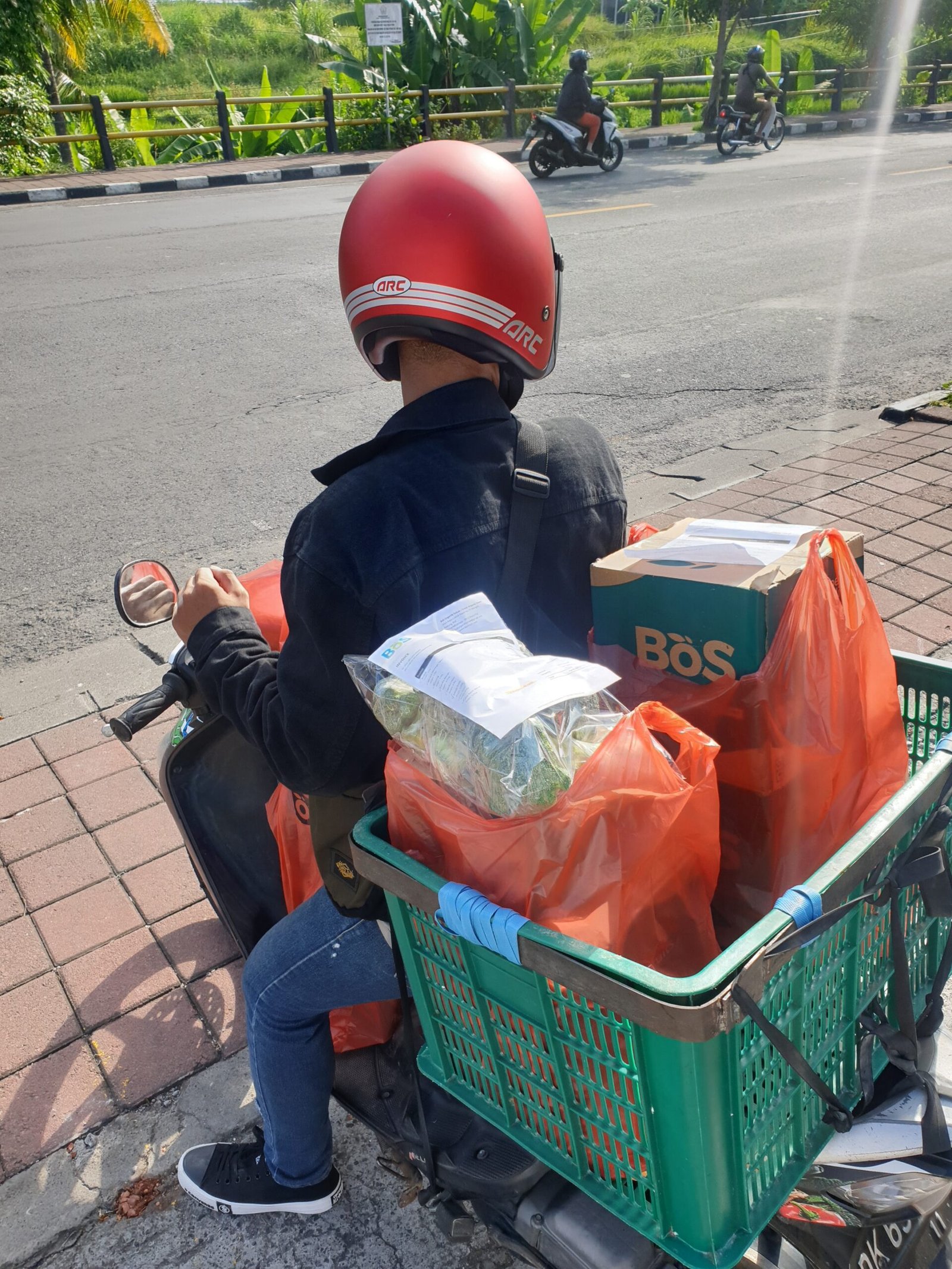 Kurir berhelm merah membawa kotak BoS dan kantong pesanan di sepeda motor, dilihat dari belakang.